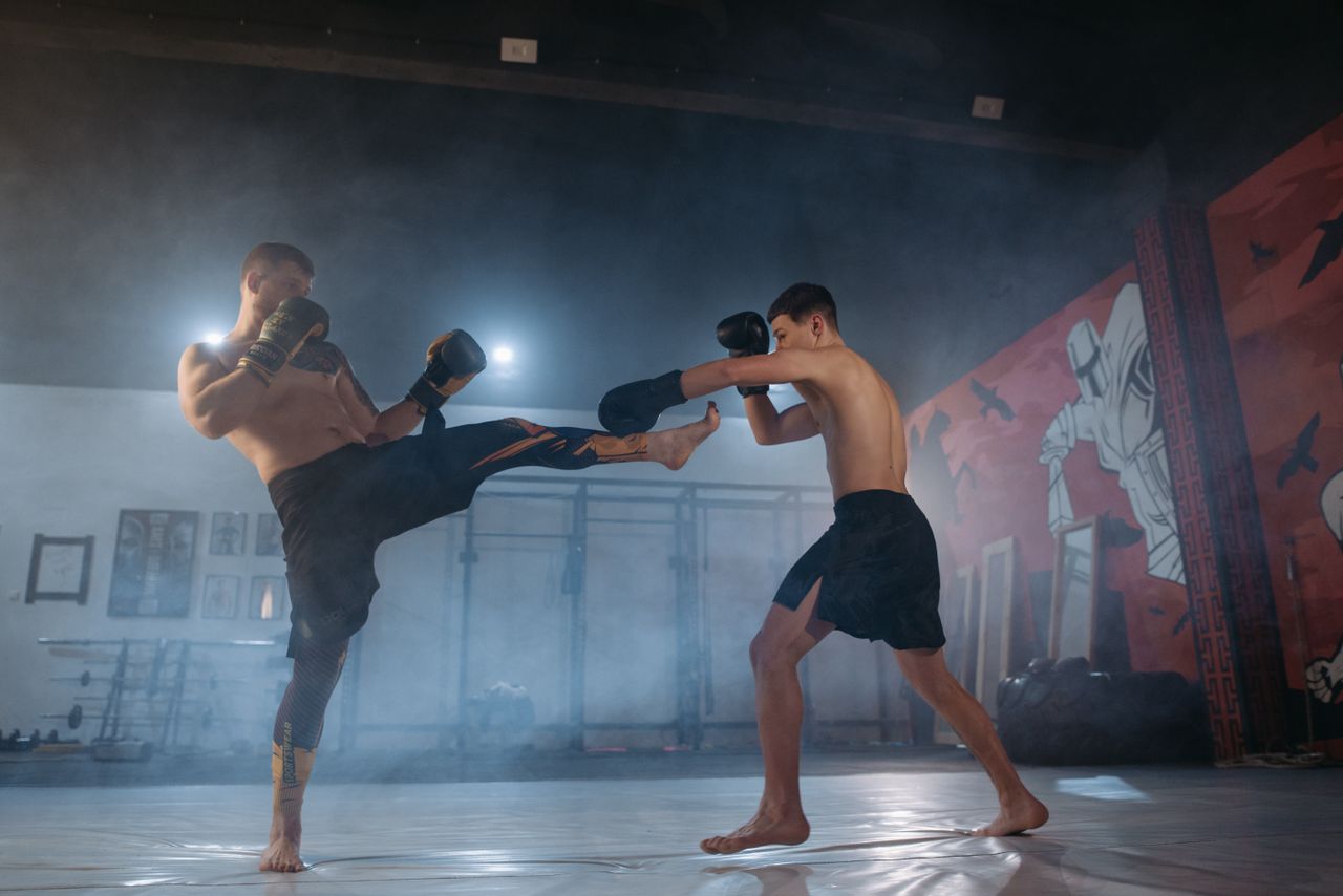 Two male fighters engage in an intense martial arts sparring session indoors with dramatic lighting.