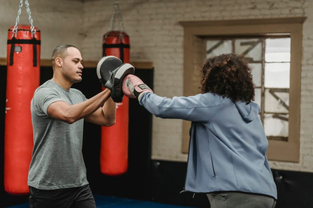 Ethnic male instructor with punch mitt training with anonymous sportswoman in boxing glove during workout in gymnasium